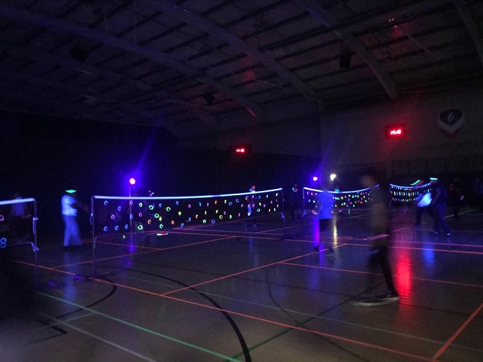 Students participating in UV Badminton in a sports hall. The students have brightly coloured clothes on and the nets have glow sticks and vibrant tape on them, which reflects off the UV light. The sports hall is dark as the main lights are turned off to give maximum effect.
