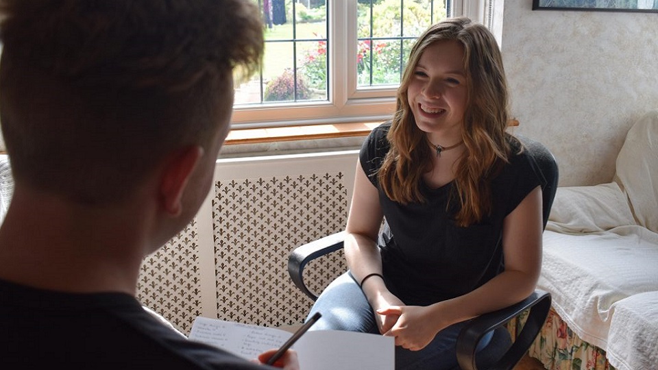 Lauren sitting on a chair chatting to her brother who is writing notes in a notepad. 