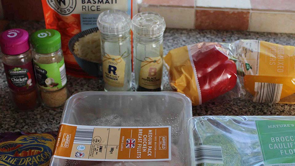 Photo of some ingredients to make a meal. There is chicken, peppers, rice, broccoli and cauliflower alongside herbs and spices.