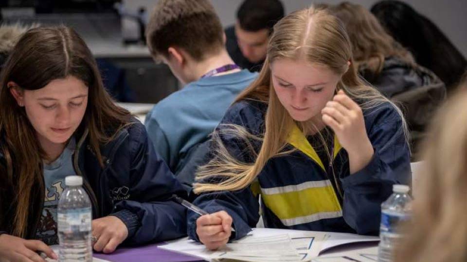 Two students looking down to a paper workbook on a table filling it in with a pen.