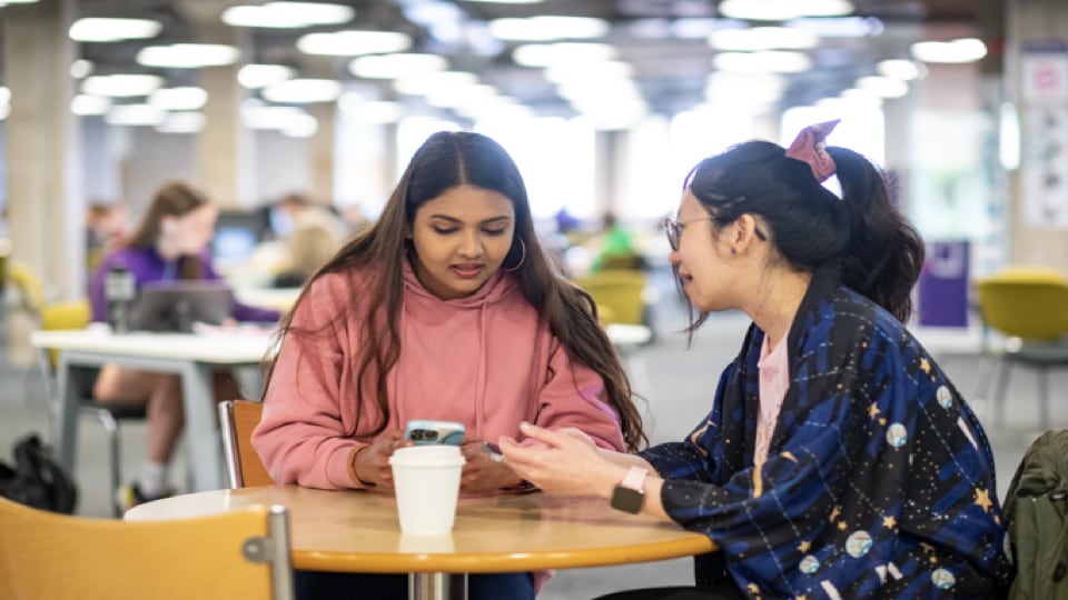 Two students sit together at a round wooden table in a brightly lit, modern university library.