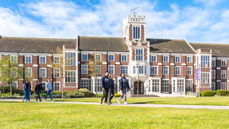 A wide, bright shot of a large, multi-story brick university building with white stone trim and a central clock tower under a clear blue sky. Two separate groups of students are walking along a paved path in the foreground across a lush green lawn.