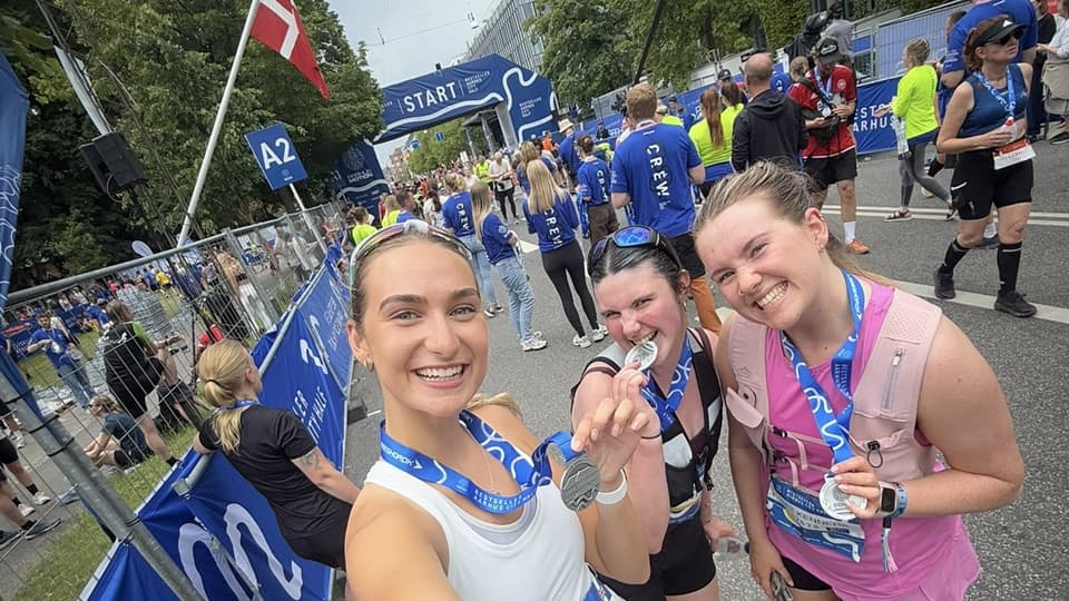 BSc Management student, Izzy Sims holding a running race medal with two friends at the finish line.