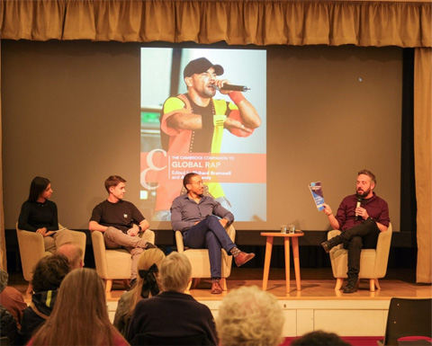 Four people sit on stage in discussion, with one holding a booklet. A projected image of a rapper and a book cover is displayed behind them. Audience members are visible in the foreground.
