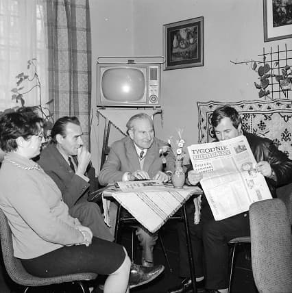 a group of people in Poland sitting around a table, one is reading a newspaper a television is in the background