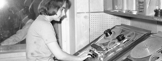 a woman operating vintage reel-to-reel audio recording equipment in a studio.