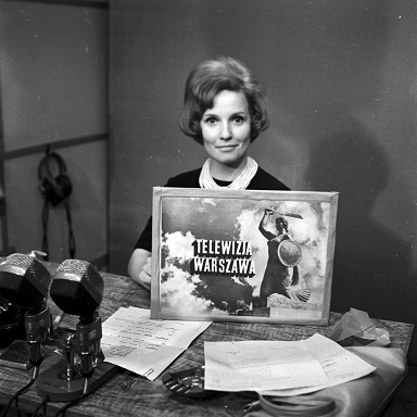 Polish TV announcer sitting at a desk holding a sign with text, Telewizja Warszawa