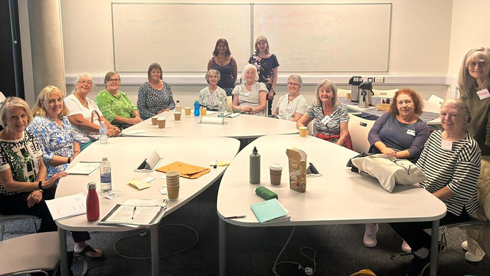 A group of women sitting in a line around a large table, three women standing in the background