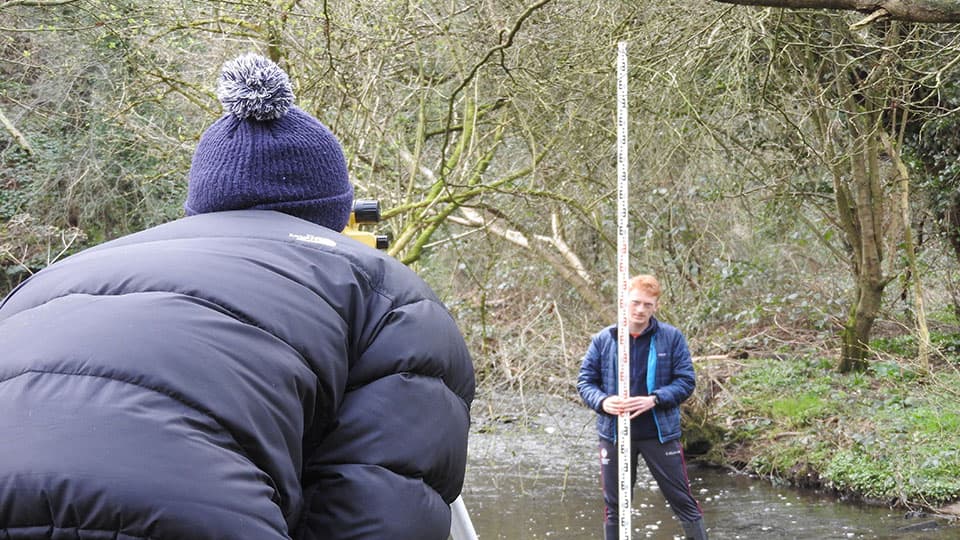 a student on the bank and another student in the stream using surveying equipment