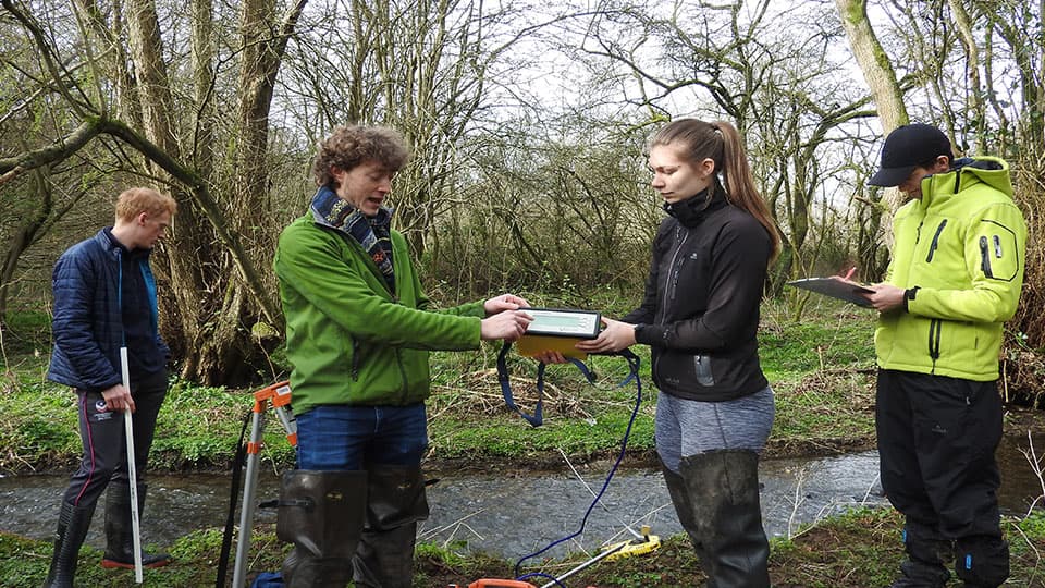 four students standing next to a stream using monitoring and measuring equipment