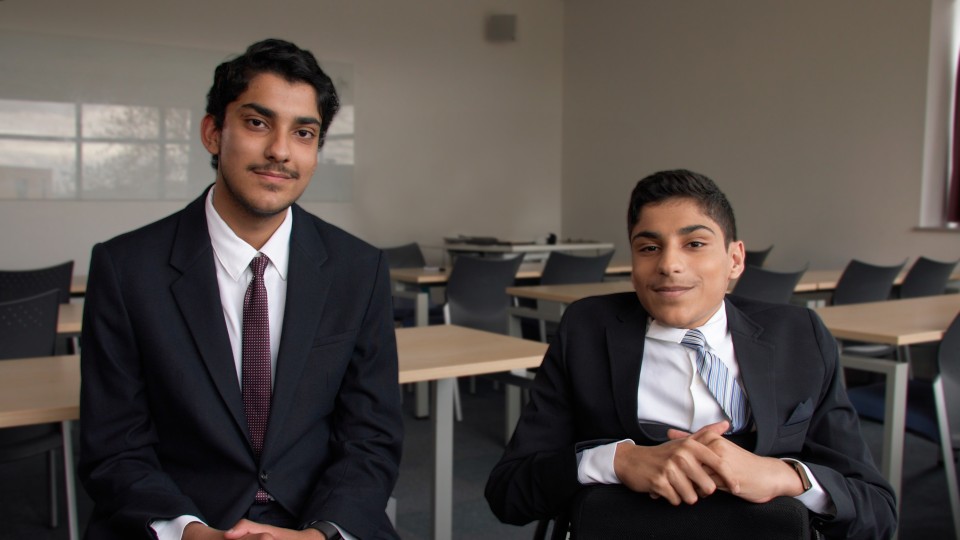 Two students sat together wearing suits with empty tables and chairs in the background.