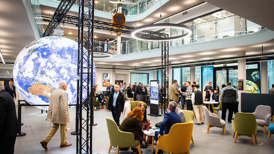 A well-lit industrial-style room with people stood or sat down on chairs networking, with a large Earth globe seen in the back of the room