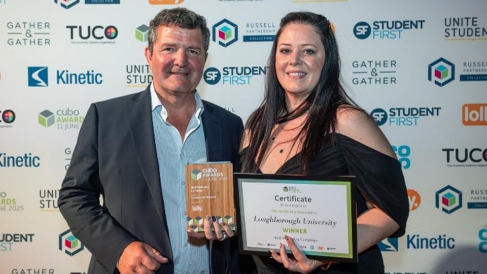 A man and a woman stood together holding a certificate and award plaque, smiling at the camera
