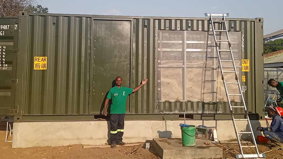 A person in a green shirt standing in front of a large green shipping container with tools and buckets on the ground.