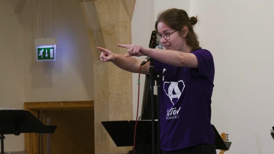 Daisy Newton wearing an Action t-shirt and pointing forwards whilst speaking into a microphone.