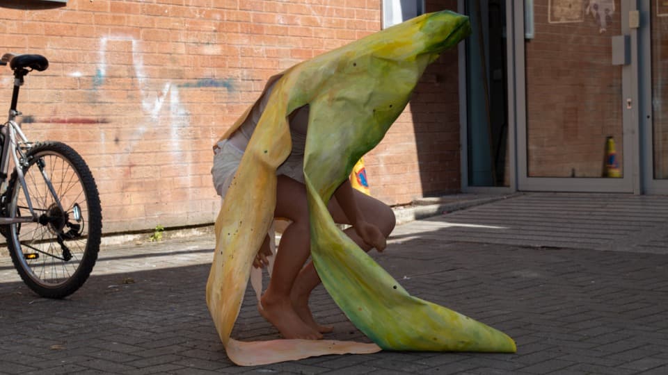 Still image from Emily McIntosh's performance 'Peel my Skin', showing her wearing a banana peel costume and crouching down in her back garden