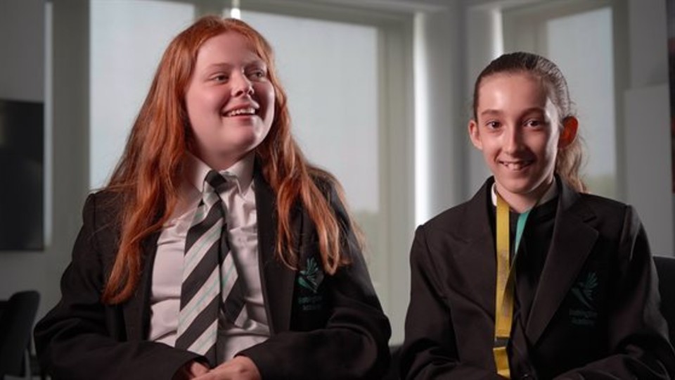 Two young people wearing school uniforms sat next to each other and smiling.