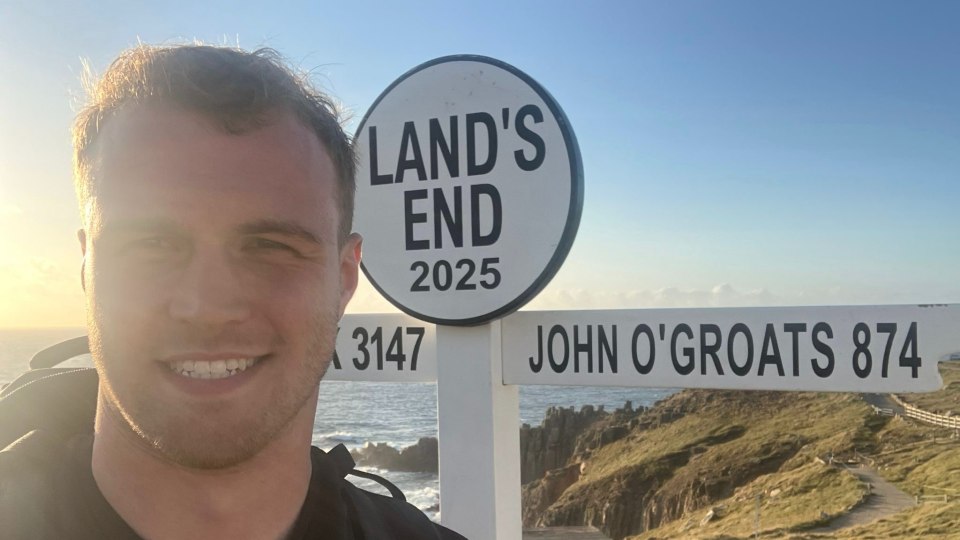 Hal Hodkinson smiling in front of a sign reading 'Land's End 2025 and John O'Groats 874 with a landscape in the background.