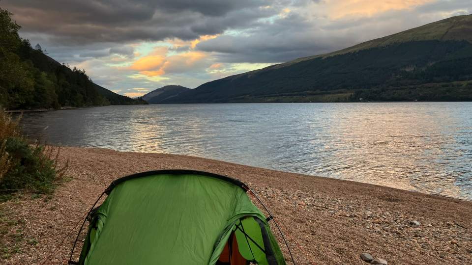 A green tent pitched opposite a lake with a view of a landscape.