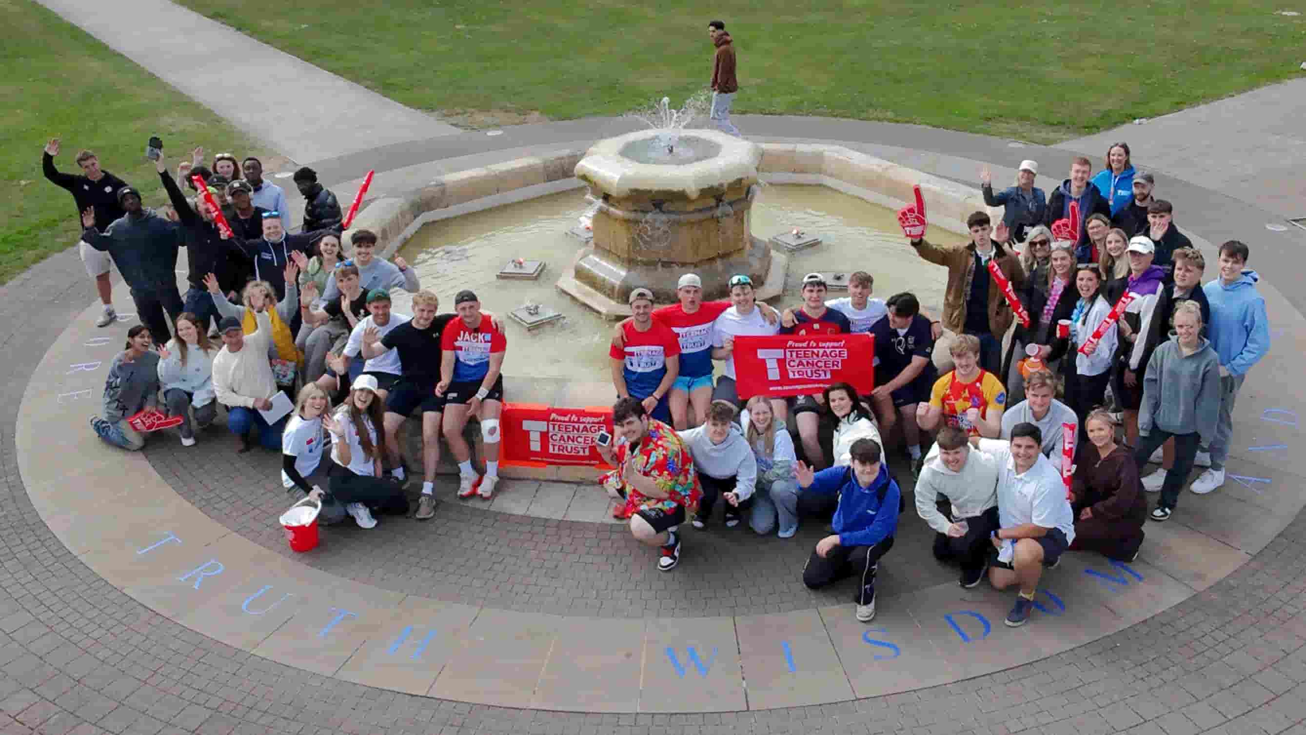 A group of people celebrating at Hazlerigg fountain, some holding Teenage Cancer Trust flags.