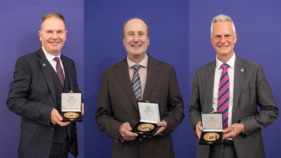 Photo of three men wearing suits holding their University Medal stood in front of a purple wall