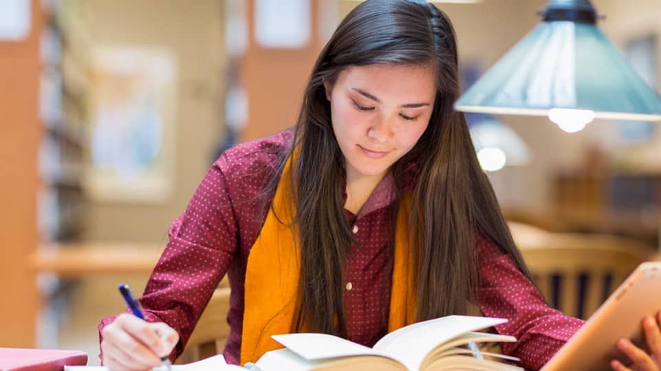 A student reading a book in a library setting and taking notes