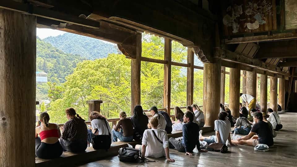 A group of students sitting on the floor under a wooden archway looking out at a view of trees.