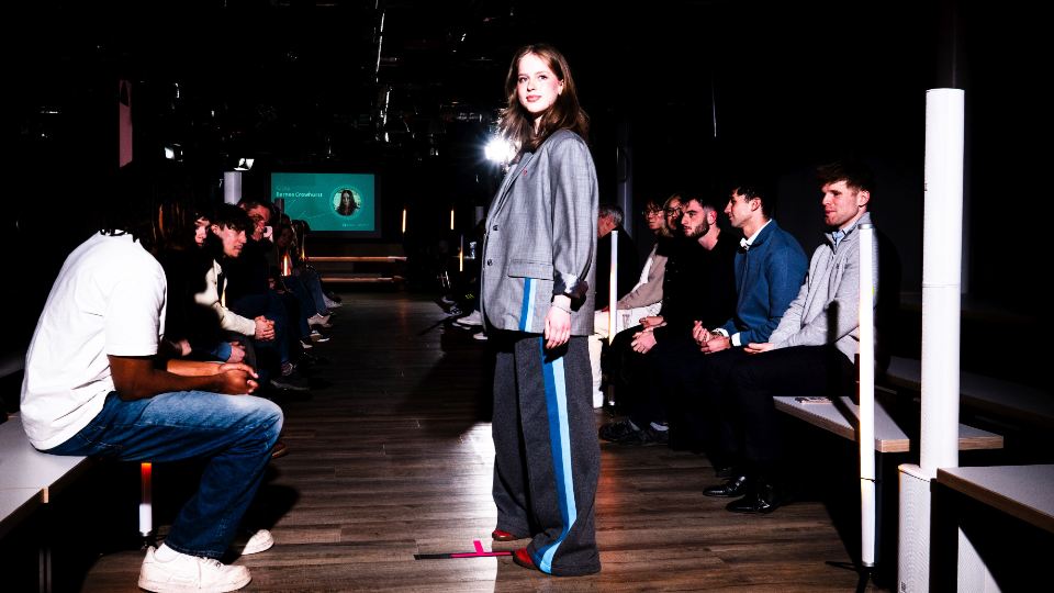 A model standing at the end of a catwalk wearing a grey blazer with blue stripes.