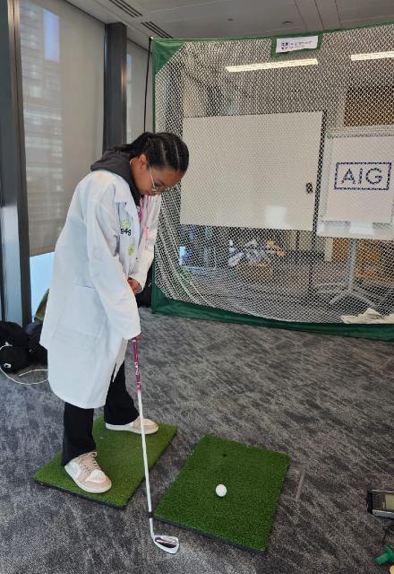 A girl wearing a lab coat aiming a golf club towards a golf ball on a patch of astroturf.