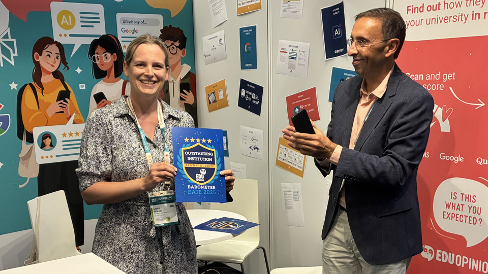 A smiling representative from Loughborough University holding a certificate for Most Outstanding Institution 2025. They are stood alongside the award issuer, who is smiling and clapping/