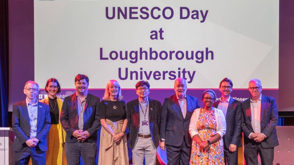 Photo of the UNESCO Chairs, senior staff from Loughborough, and representatives of UNESCO UK stood together on a stage smiling at the camera