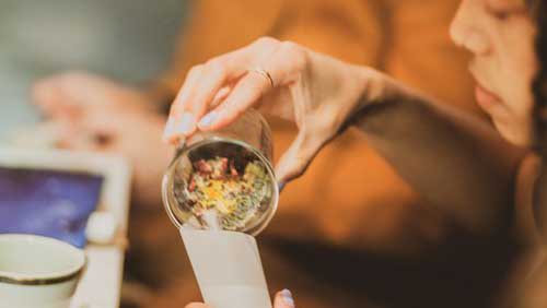 A person pouring herbs and petals from a plastic cup into a paper envelope.