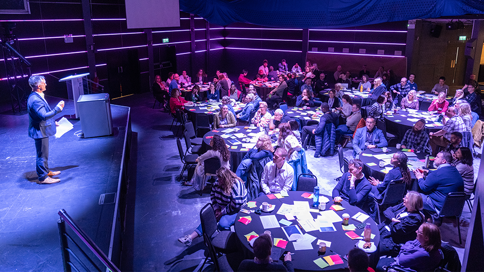 Professor Sam Grogan stood on a stage speaking in front of a large group of staff sat at roundtables