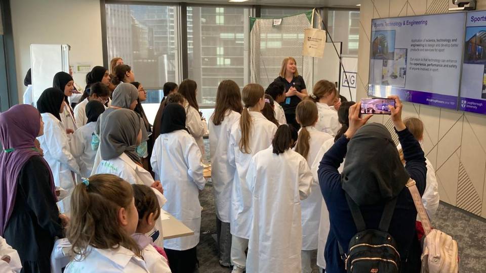 A student presenting to a group of young girls wearing white lab coats.