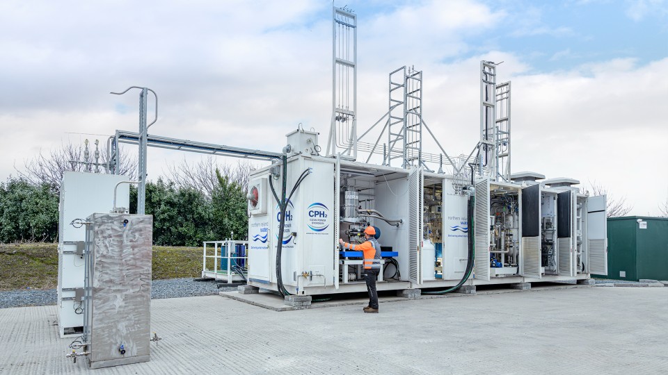 Worker operating modular hydrogen production units outdoors with pipes and tall metal structures.