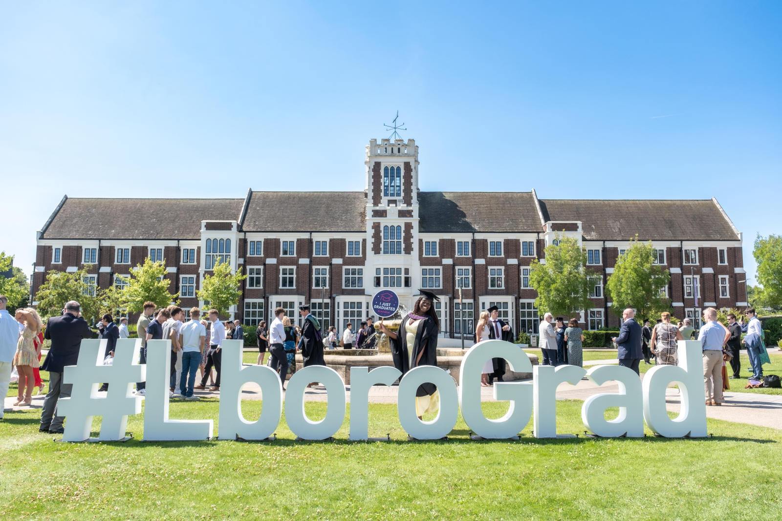 Photo of the Hazlerigg Building and lawn on a sunny day with big white letter signage spelling out ''#LboroGrad' on the lawn, with people behind it wearing graduation gowns