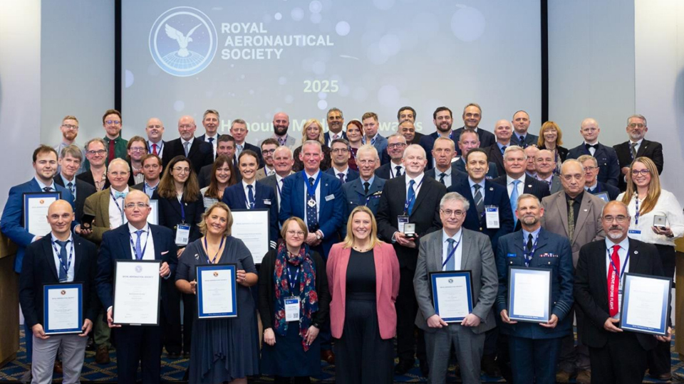Group photo at an awards ceremony for the Royal Aeronautical Society 2025, with attendees holding framed certificates and standing in front of a large screen.