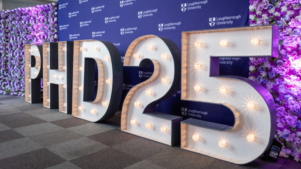A 3D sign 'PHD 2025' standing in front of a purple backdrop with the Loughborough University logo on it.