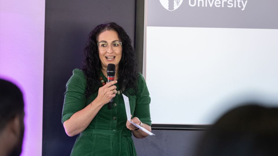 Photograph of a woman holding a mic and speaking, stood next to a screen