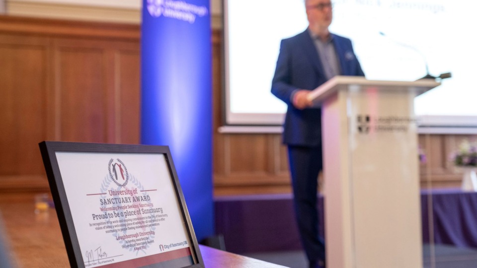 The framed certificate for Loughborough's University of Sanctuary award, with the Vice-Chancellor stood on a lectern making a speech